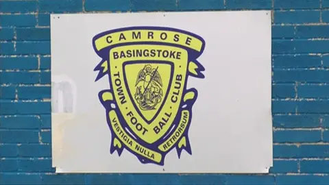 The Basingstoke Town logo is yellow and blue, and is printed on a white sign. The sign is on a blue wall