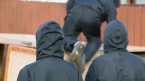 Three people wearing black hoodies and facing away from the camera. One is climbing over an object in front of them.