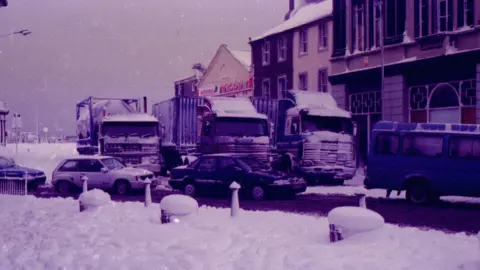 Cars and lorries queuing in Whitehaven town centre as the town is covered in snow. The tops of three bollards are barely visible due to the heavy snow covering them.