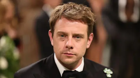 A close-up image of Jonathan Anderson pictured at the Met Gala in 2024. He has short brown hair and has a neutral expression on his face. He is wearing a black suit jacket with a shamrock broach on his shoulder.