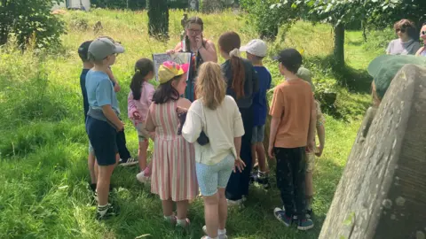 BBC A group of children are standing close to a large gravestone. They are in a green space with a large number of trees. 