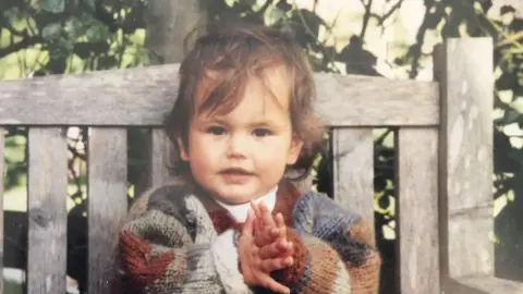 Judy Young A photograph of a young child with wispy brown hair. She is sat on a wooden bench in a colourful cardigan. 