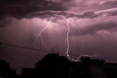 A purple sky as thunder is strikes above a silhouette of houses and trees.