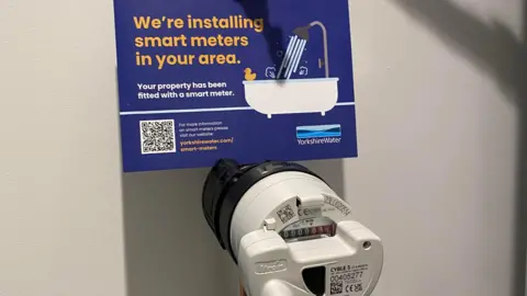 Yorkshire Water A smart water meter installed on an internal copper stop tap, it is a white plastic circular device with a meter reading on it. There is a Yorkshire Water card above which reads, we are installing smart meters in your area.