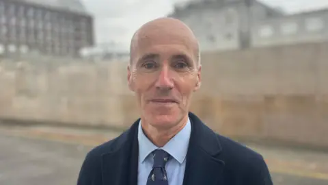 BBC Deputy Renouf is a man with a bald head, staring into the camera. He is wearing a navy coat, with a blue shirt and navy tie. He is standing in front of a wall with buildings out-of-focus behind him.