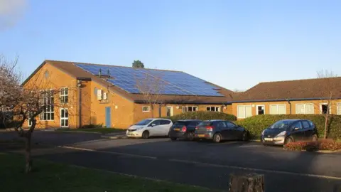 Jonathan Thacker/Geograph Outside Wittering Primary School. It is a one-storey building with solar panels on the roof. There are four cars parked outside.