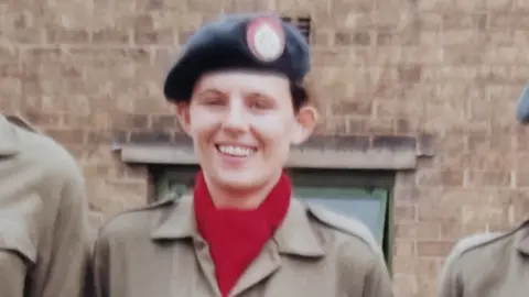 Karen Usher A slightly blurry picture taken in the 90s, shows a young girl in an RAF beret, and a red scarf. She is wearing what looks like khaki overalls.