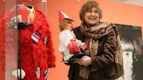 Showtown Lady Anne Dodd has a brown fur jacket, a patterned brown scarf and holds a Diddyman. In the background is a large red fur jacket with two tickling sticks in its pockets.