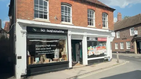 A double-fronted detached shop building on the corner of a street. The upper floor is built from red bricks with large windows. The ground floor features two shops, either side of a central doorway. The one on the left is called So Sustainable, the one of the right is vacant with a 'for sale' sign in the window.
