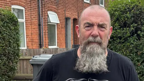 Jaimielee Rendall/BBC A man with a beard and black t-shirt holds onto a fence and looks at the camera 