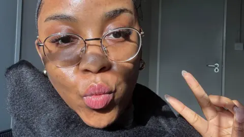 Rosemary Nwodoh is seen in a selfie taken at work. The closeup photograph shows her with a scarf around her neck and wearing glasses. She is sitting in a grey office with a door behind her.