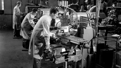 Walter Nurnberg/Getty Black and white photo showing young men working at lathes, wearing long white coats. 
