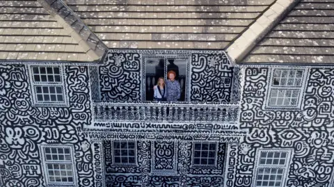 Reuters Mr Doodle is standing on the balcony of his home, with his wife
His house is covered in black cartoonish drawings against a white background
His roof is the only part that has not drawn on.