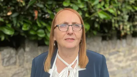 A woman with shoulder length red hair stands in a street - she is wearing a blue jacket and white shirt and glasses, standing in front of a stone wall with foliage growing on top of it