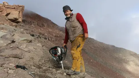 Prof John Marshall stands on a steep rocky slope and holds a large drill. He is "drilling the End Devonian mass extinction at 1000m on Celsius Bjerg, East Greenland", according to the University of Southampton.