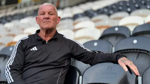 BBC Newsnight Windass, with short grey hair, is sitting on a black seat in Hull City's football stadium. He is looking directly into the camera lens and wearing a black sports top with white stripes down the side.