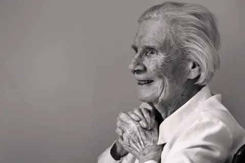 A black and white photo of an elderly lady sitting in a chair against a plain wall, smiling with her hands clasped as she looks out of the frame.