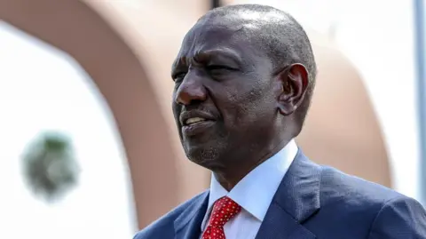 A head and shoulders shot of President William Ruto wearing a blue shirt and red tie.