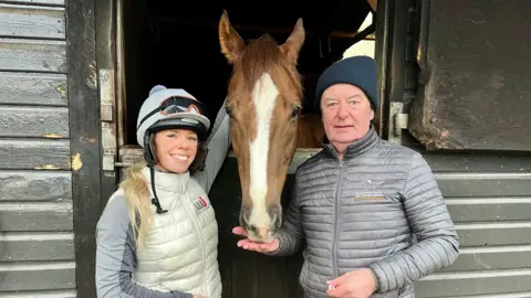 DAVE EDWARDS/BBC A woman and a man stand either side of a brown racehorse at a stables.