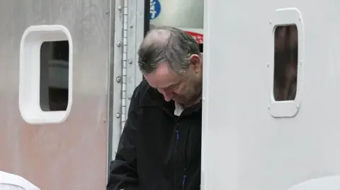 A man with thinning grey hair wearing a black zip jacket. He is facing down while walking out the door of a grey vehicle with small windows either side of the door