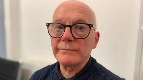 Paul O'Neill is bald with grey hair at the sides and is wearing black-framed glasses and a dark jumper and shirt