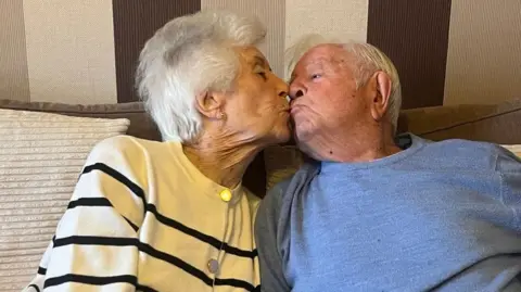 A picture of Ted and Ellen mimicking a kiss. Both have white hair and are sitting on a sofa. Ted is wearing a blue jumper and Ellen is wearing a striped cardigan. 