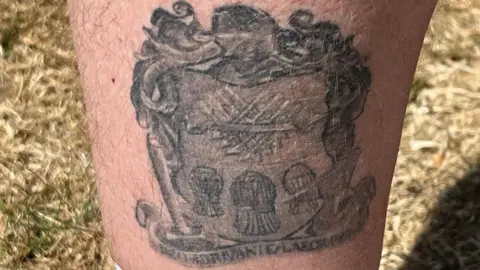 A crest with a helter skelter and some latin characters. There is limited contrast between the colours making the words and objects quite difficult to make out without knowing what each element is.