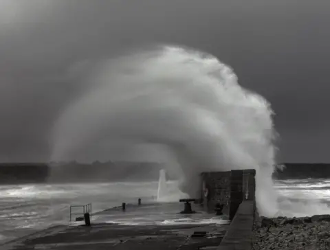 Angela Pritchard A black and white photograph of a large wave crashing against a sea wall