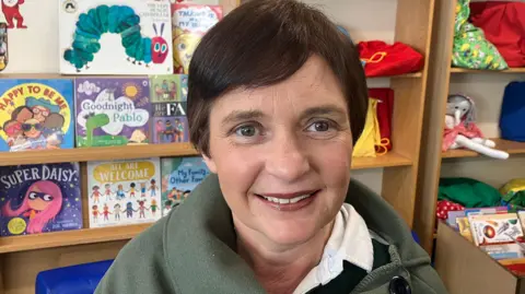 BBC Tracy Ratstone smiling on camera. He has short brown hair and is wearing a green top. He is behind books and toys behind it.