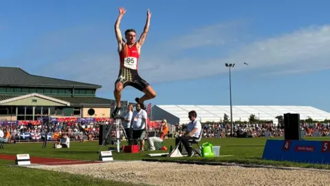 Manx Radio Sport Regan Corrin jumps high with both arms in the air over a strip of sand in the long jump competition. He wears a red and black lycra jumpsuit. 