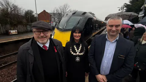 Jim Scott/BBC Grahame Morris MP, left, and wearing a cap stands with Seaham's Mayor Margaret Levitt - wearing all black and with the chain office over her shoulders - and Chris Brandon from Grand Central in a dark suit and tieless light blue shirt. They are standing on the platform at Seaham where the yellow fronted Grand Central train has arrived. In the background, many people are holding up umbrellas.