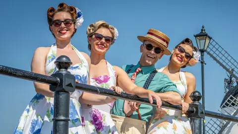 Andy Bell Images Three women and a man in vintage 1940s-style summer clothing and sunglasses lean against a black railings. The women are wearing floral dresses, while the man wears a striped straw boater hat, shirt and braces. A traditional windmill is visible in the background against a clear blue sky.