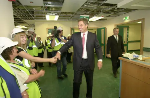PA Media A man in a suit with a white shirt and pink tie is smiling and shaking the hands of a row of workers in hard hats and hi viz vests