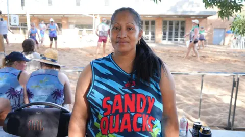 A woman, with long black hair and wearing a sports singlet with the words Sandy Quacks, in front of a beach volleyball court