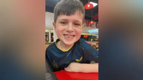 Archie Benwell, a seven-year-old boy with brown hair, smiles at the camera. He is wearing a Newcastle football shirt.