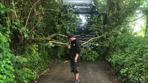 A front view of a lorry stuck on a narrow lane. A man wearing shorts and a black hoodie and baseball cap is stood in front of the lorry.