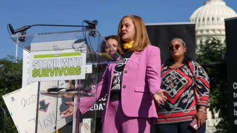 Reuters Liz Stein, who was a victim of late financier Jeffrey Epstein, speaks on the day of a rally in support of Epstein's victims, on Capitol Hill in Washington, DC, in September 2025. Liz is wearing a pink suir and standing in front of a podium with the word stand with survivors on a sign. 