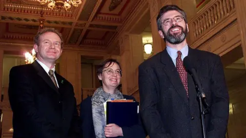 Gerry Adams, Martin McGuinness and Bairbre de Brun standing together smiling. Gerry Adams has a microphone in front of him. He is wearing glasses, a suit, light blue shirt and red patterned tie. Bairbre de Brun is holding a file and is wearing a grey patterned scarf, navy jacket and glasses. Martin McGuinness has a black suit on with a white shirt and gold patterned tie.