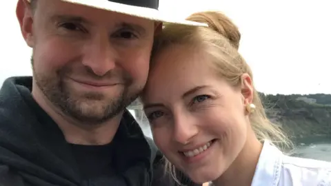 Christopher Jones is on the left and is wearing a black T-shirt, black jumper and a white hat. He has a short brown beard and moustache. Standing next to him on the right is his wife Paula, who has blonde hair in an updo and is wearing a white shirt.