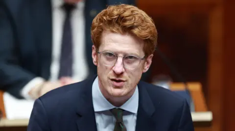 PA Media Matthew O'Toole wearing a blue suit, white shirt and green tie. He has red hair.