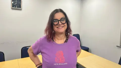 Jessica Bayley/ BBC A lady with shoulder length dyed red hair, wearing glasses with black thick frames stands facing towards the camera. She is wearing a purple t-shirt which says 'New Zealand' on it, she also has bracelets on her wrist. There is a clock on the wall in the background.