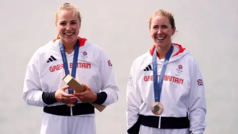 PA Media Two women dressed in white tracksuits with Great Britain across the chest in red lettering holding a bronze trophy while wearing bronze medals