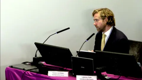 Lampard Inquiry Thomas Coke-Smyth is sitting in the inquiry room at Arundel House in London. He is sitting behind a thin microphone in front of a purple table and a series of laptops. He is wearing a black suit with a pale yellow patterned tie and a white shirt. He is addressing Cawston during the inquest