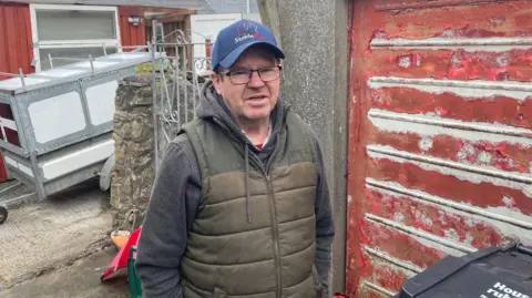 Man standing by a bin in a back lane in Penzance  wearing a cap