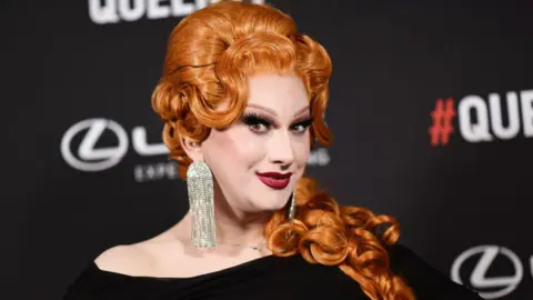 Getty Images Drag Queen Jinkx Monsoon - in glamorous Hollywood make-up and red lips, with a bright ginger curled wig and huge diamante earrings. She is smiling at the camera while attending the Queerties event in 2024.