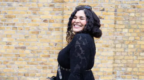 Gemma Devonish, a woman with long, dark hair, smiling and wearing a black dress 