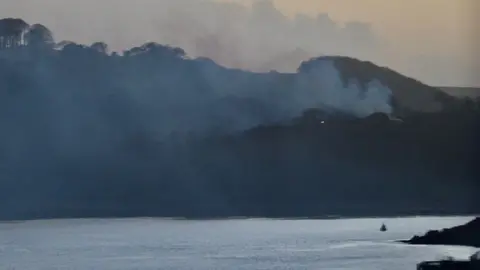 Smoke is rising from a hill seen from across a river