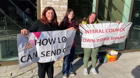 Tom Walker/BBC Two women and a girl stand side by side. The two women are holding two different signs which read, "how lo can send go?" and "Wirral Council own your shame". 