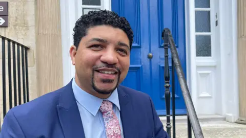 Glenroy Bell is standing to the left of the image looking towards the camera smiling. He has short brown hair, and is wearing a navy suit, blue shirt, and a pink floral tie. He is standing in front of a blue door.