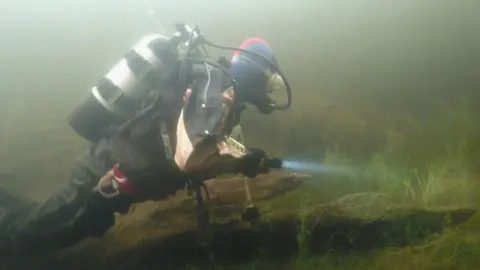 A diver with an oxygen tank and mask uses a torch while exploring the Loch Achilty crannog.